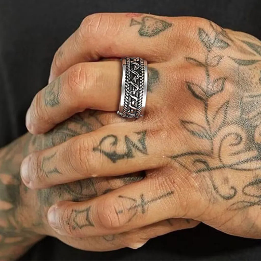 Gros plan sur deux mains tatouées, bague Skadi en acier inoxydable au motif torsadé central, mise en scène virile et stylée.