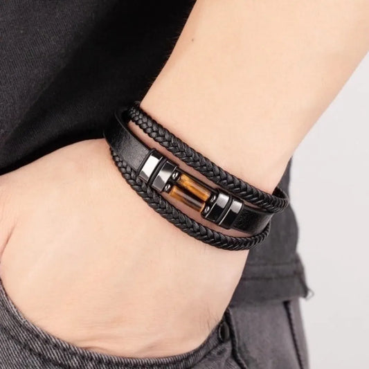 Bracelet pour homme en cuir tressé noir, porté élégamment au poignet gauche. Le bijou est orné d'une pierre œil de tigre jaune lumineux, mettant en valeur son design sophistiqué et son confort, idéal pour un usage quotidien.