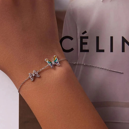 Gros plan sur le bracelet Papilla en argenté, porté sur un poignet féminin devant une affiche CÉLINE, mettant en valeur ses deux papillons finement travaillés.