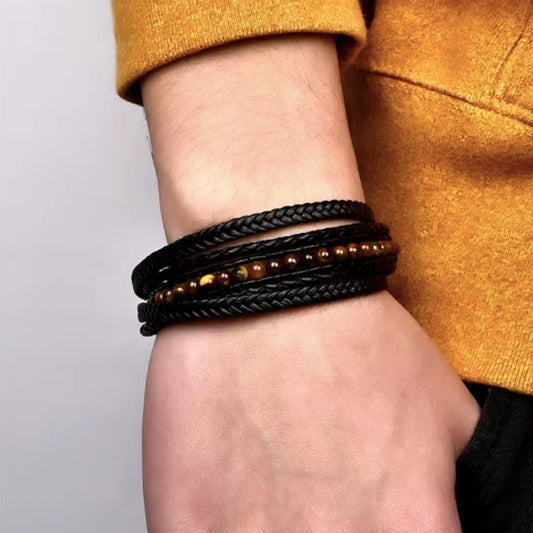 Bracelet multitours en cuir noir tressé, orné de perles naturelles œil de tigre, porté sur le poignet d’un homme. Le bracelet s’associe à un pull jaune moutarde, créant un contraste élégant et chaleureux. L’arrière-plan est neutre, mettant en avant les détails et la texture du cuir, ainsi que la brillance subtile des perles œil de tigre. Un accessoire moderne et sophistiqué pour un style masculin affirmé.