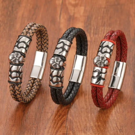 Trois bracelets en cuir tressé, côte à côte sur une table en bois au ton chaleureux. Chaque bracelet présente un fermoir magnétique en acier inoxydable argenté, avec des motifs gravés audacieux au centre, dont une tête de mort emblématique. Les bracelets affichent chacun une couleur distincte : beige, noir et rouge. Leur design harmonieux et leur disposition en arc mettent en valeur leur élégance et leur caractère intemporel, sans arrière-plan supplémentaire pour une présentation minimaliste et percutante.