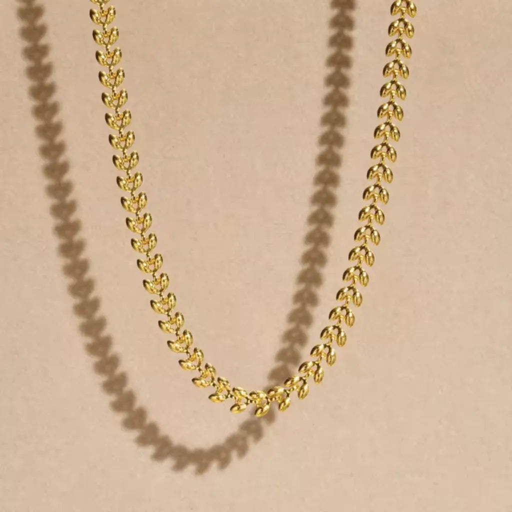 Collier doré en double chaînage, ombre portée sur fond beige, effet visuel raffiné pour souligner la texture feuillage et le plaquage or 18K.