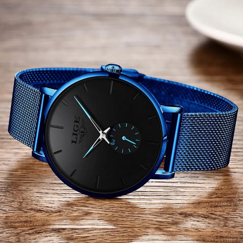 Photo d'une montre Top Casual minimaliste reposant sur une table en bois, son cadran, sous-cadran et ses index sont noir - les aiguilles fines et le bracelet en maille milanaise sont de couleur bleu roi