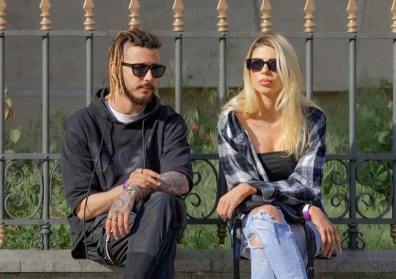 Jeunes couples assis côte à côte sur un petit muret en plein solei, les jambes et les bras croisés, ils arborent tous les deux une paire de lunettes de soleil de style voyageur de couleur noire