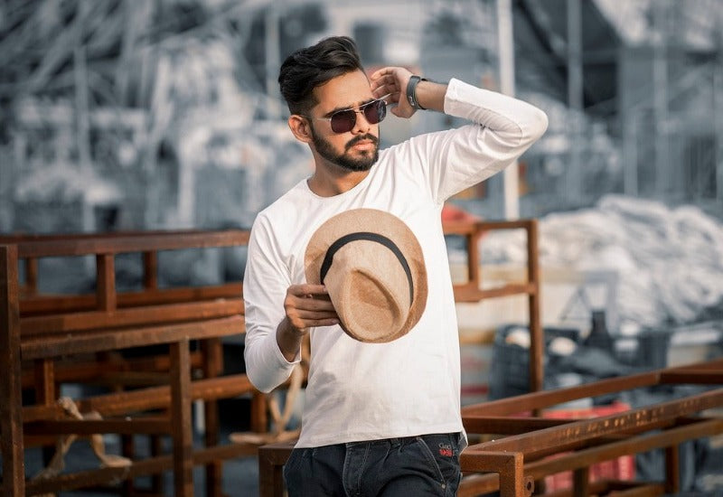 Homme barbu vêtu d'un tee-shirt blanc en manches longue tenant un chapeau à sa main droite, lunette de soleil noire style pilote sur le nez et montre bracelet cuir au poignet gauche