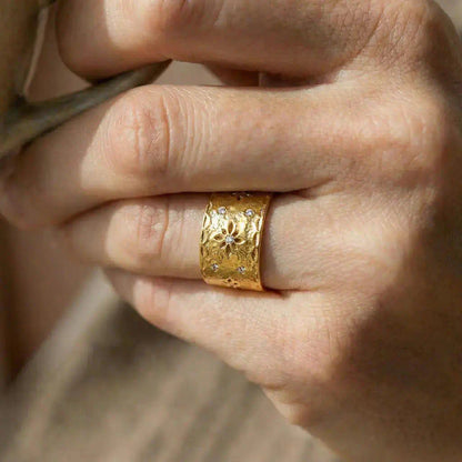 Zoom sur la bague Aura large et ajouree portee au doigt d une femme tenant une tasse montrant le motif floral central avec cristal brillant sur la surface texturee.