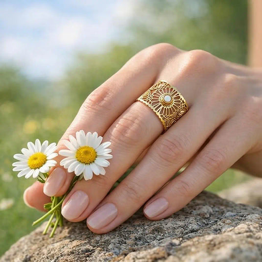 Bague Lyna ajustable acier fusion or portée main profil marguerites motif floral ajouré cabochon blanc éclatant décor naturel rocher ciel nuageux.