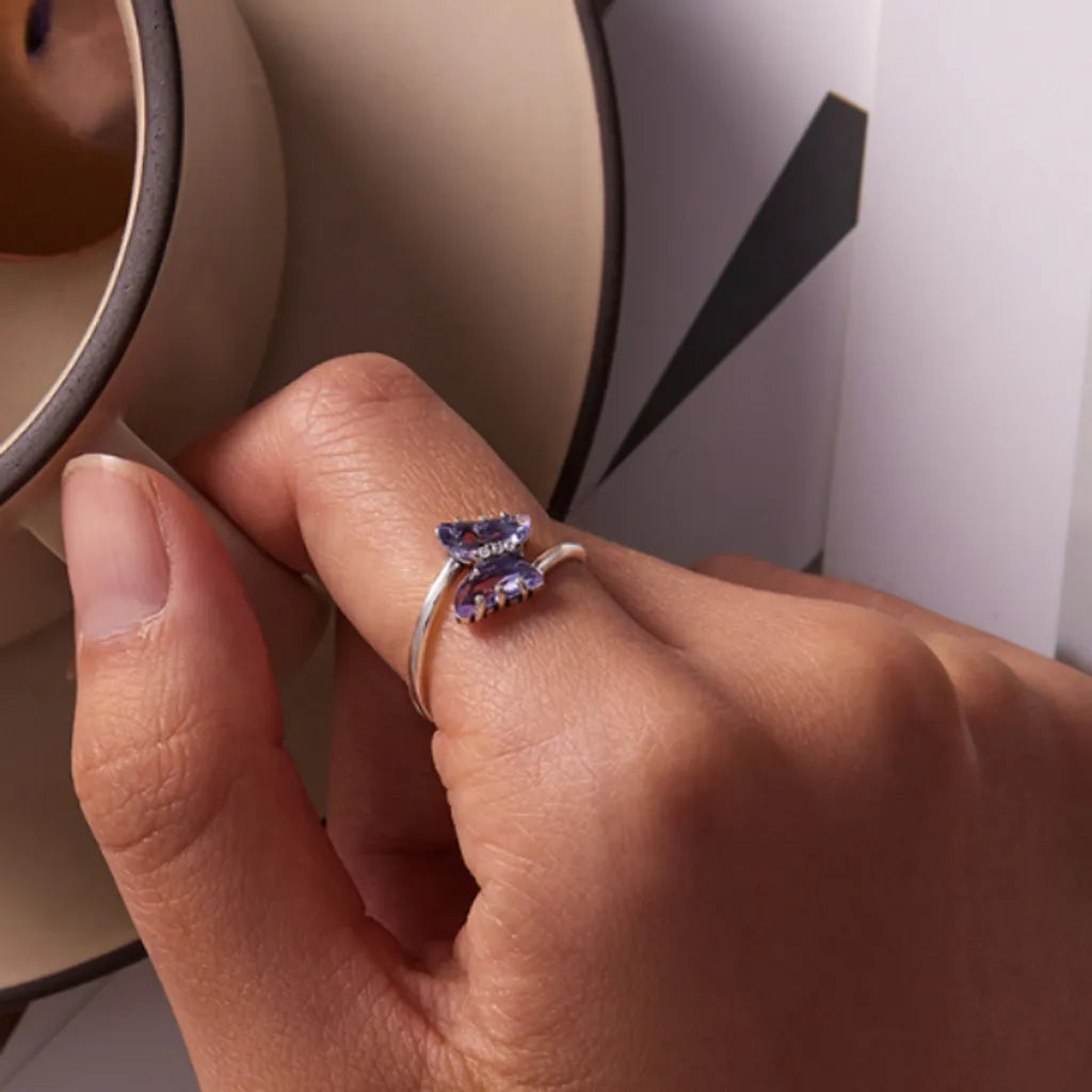 Bague papillon en argent 925 avec cristal violet et zircones, portée délicatement à la main. Le décor naturel met en valeur l’élégance du bijou avec une touche moderne, une tasse en céramique et un fond graphique subtil.