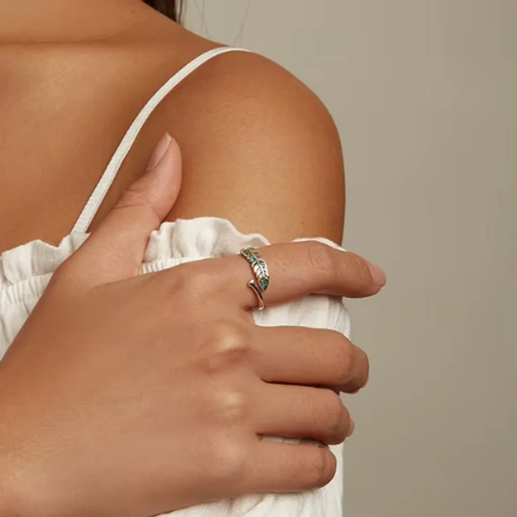 Main droite d'une femme placée sous son épaule, entourant délicatement son bras au niveau d'un top blanc à bretelles, mettant en valeur une bague plume colorée en argent 925 avec émail multicolore.