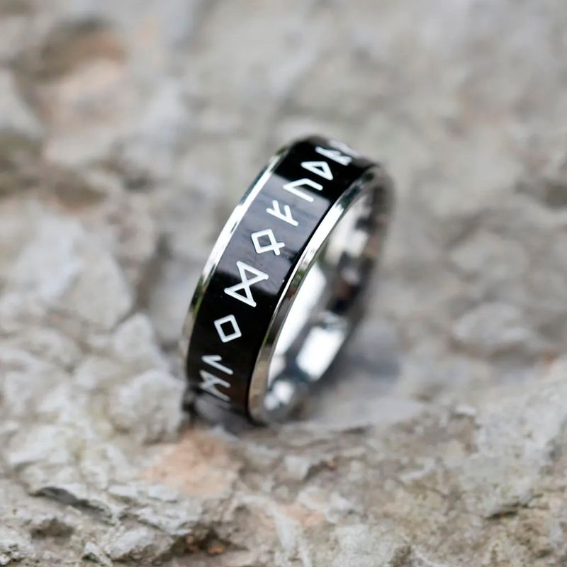 Bague Rune Viking, anneau rotatif en acier inoxydable pour homme, calé, droit et légèrement incliné sur un rocher grisâtre – coloris noir et argent.