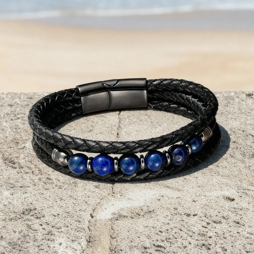 Vue aérienne du bracelet Azur déposé à plat sur un muret de pierre, révélant la souplesse du cuir multi-rangs et l'éclat du fermoir magnétique gunmetal devant un panorama marin.
