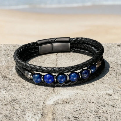 Vue aérienne du bracelet Azur déposé à plat sur un muret de pierre, révélant la souplesse du cuir multi-rangs et l'éclat du fermoir magnétique gunmetal devant un panorama marin.