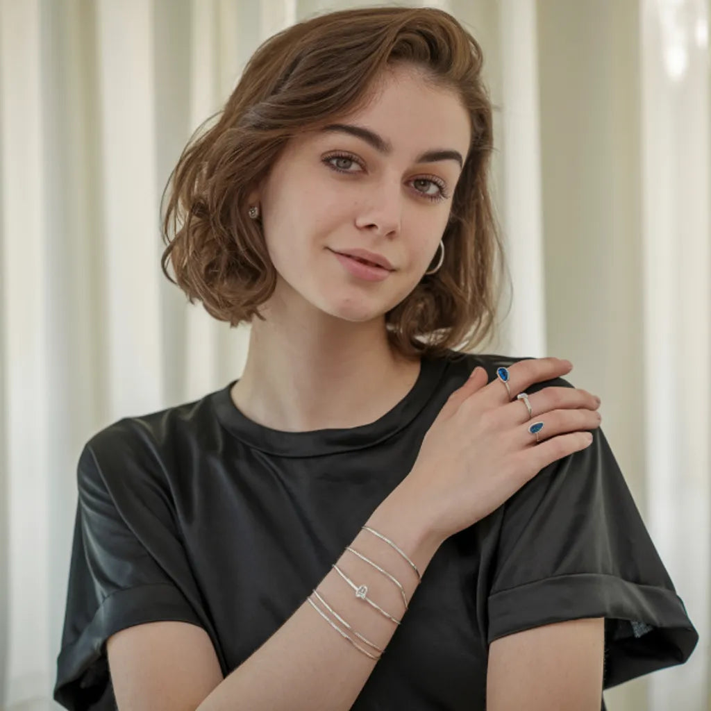 Jeune femme élégante portant des bijoux en argent 925, comprenant plusieurs bracelets fins, une bague ornée d'une pierre bleue, et une bague minimaliste. Elle pose dans un cadre lumineux avec des rideaux blancs en arrière-plan, vêtue d'un haut en satin noir, mettant en valeur l'éclat raffiné et contemporain des accessoires. L'accent est mis sur le mélange de simplicité et de sophistication des bijoux, parfaits pour un look moderne et féminin.