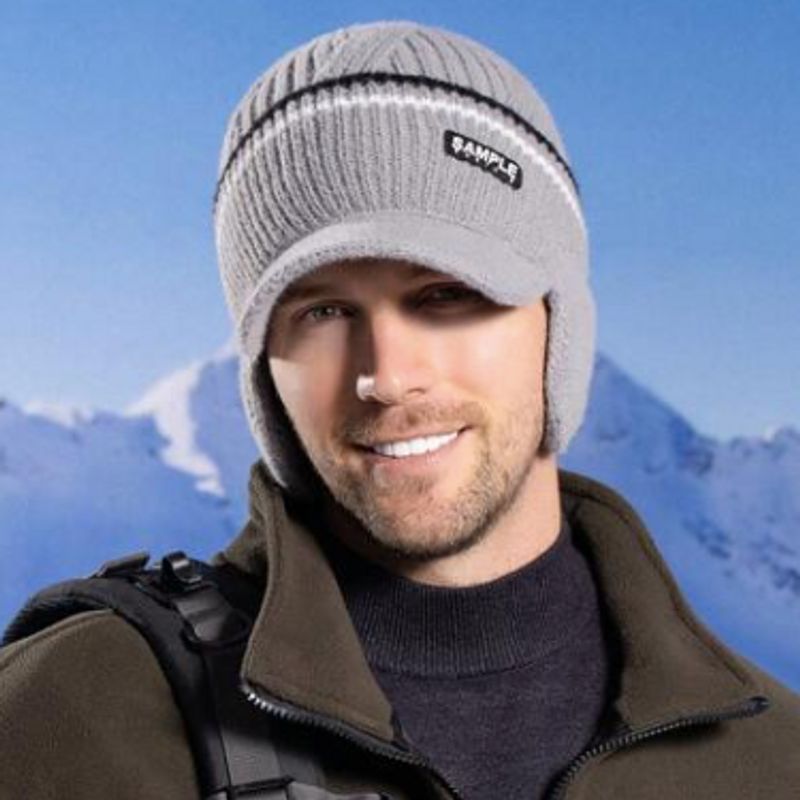 Vue sur un bonnet à visière Oslo, casquette avec cache-oreilles tricoté en laine de qualité en acrylique. Bonnet enfoncé sur la tête d'un jeune homme très souriant, prise de vue devant de splendides montagnes au sommets enneigés 