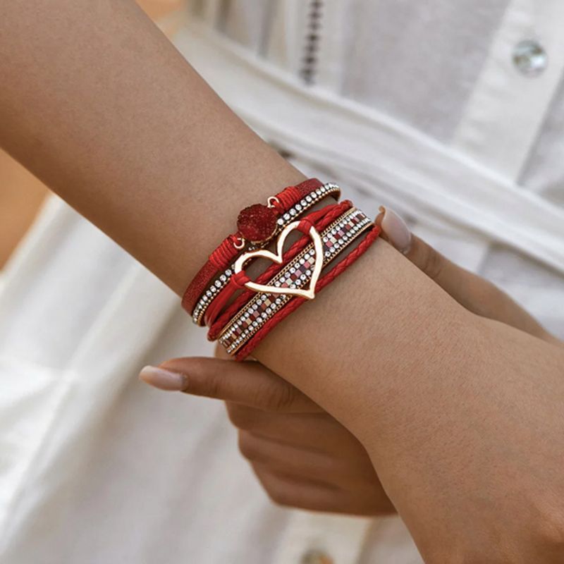 Bracelet amour de cœur, wrap bohème multilanière en cuir PU rouge présenté sur le poignet d'une femme élégante vêtu de blanc avec breloques en pierre de résine rouge et cœur ajouré doré, rivière de strass en cristal et fines chaînes en alliage de zinc. Fermoir magnétique de couleur or.