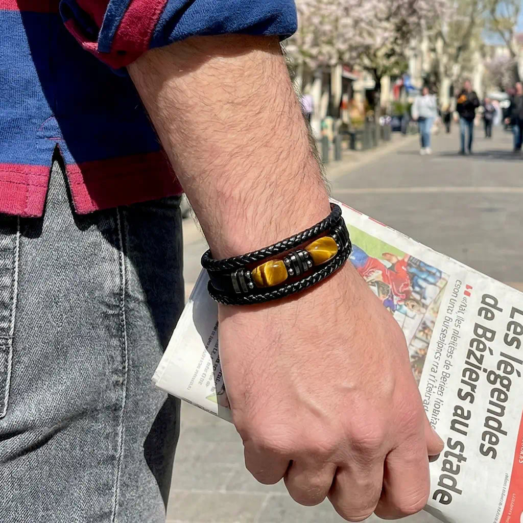 Zoom détaillé sur le bracelet Apex porté au poignet, révélant la souplesse de ses trois lanières de cuir noir et l'éclat des pierres œil de tigre alors qu'un homme tient un journal dans une rue piétonne baignée de lumière.