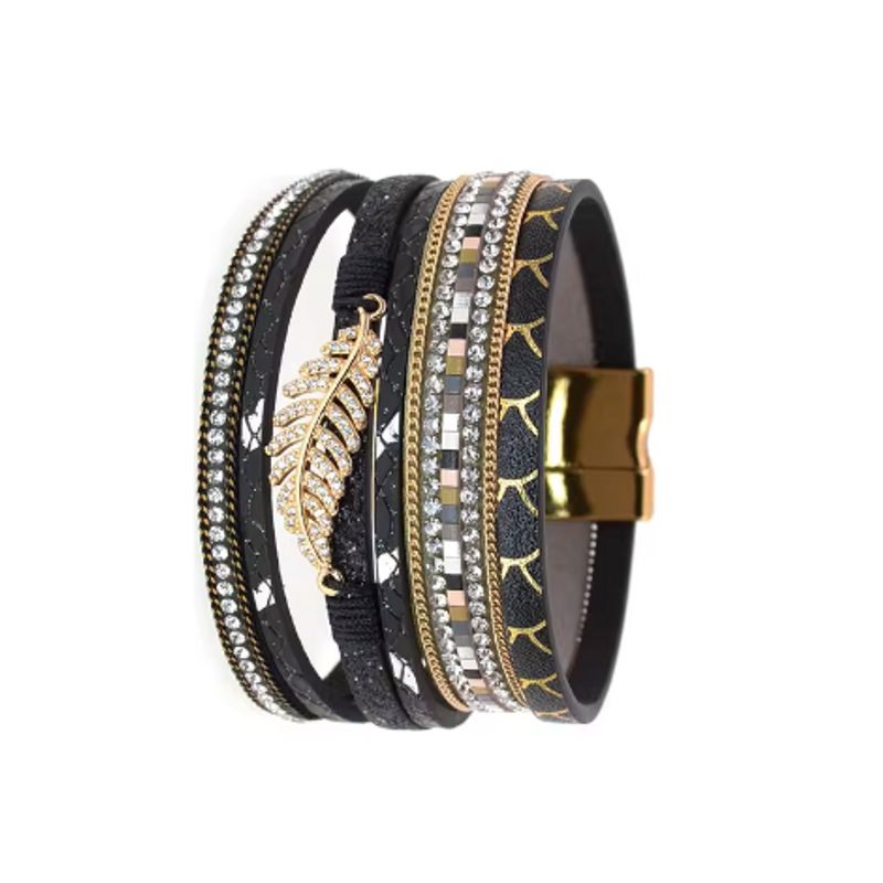 Bracelet bohème chic présenté dressé, wrap multilanière en cuir PU avec breloque feuille de fougère dorée et ornée de strass blanc en cristal, son fermoir magnétique est de couleur or. Coloris noir.