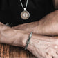 Bracelet avec chaîne à maillons vénitiens de 6 mm en acier inoxydable de couleur argent, son fermoir et un tube clip à ressort - présenté autour d'un poignet d'homme ayant les bras croisés et affichant un superbe collier pendentif rond