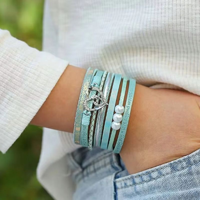 Bracelet cœur bohème argenté avec perles pour femme, wrap bohème mantras en cuir PU de couleur bleu ciel présentée autour d'un poignet féminin, la main dans la poche d'un pantalon en jean bleu délavé.