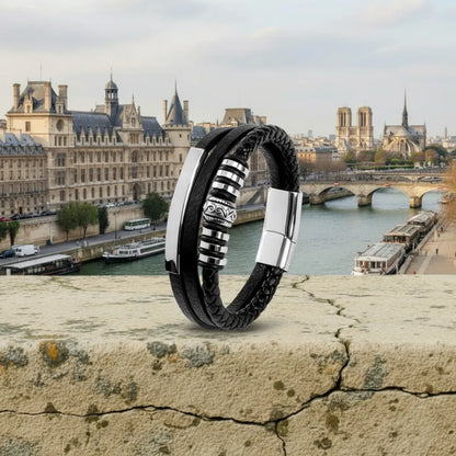Ambiance parisienne. Le bracelet Corax multi-rangs cuir noir dressé sur une rambarde de pierre, avec la Conciergerie en arrière-plan. La frise gothique et le fermoir brillent sous un ciel automnal.