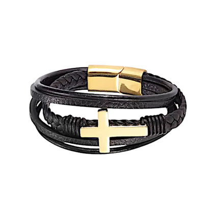 Le bracelet en cuir noir avec sa croix latine dorée et son fermoir assorti, posé à plat sur fond blanc.