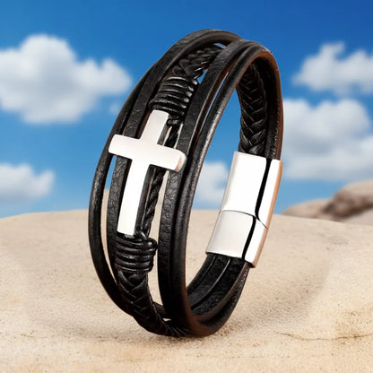 Vue 3/4 du bracelet en cuir multi-rangs dressé sur un rocher. Le fermoir et la croix latine sont mis en valeur sur fond de ciel bleu.