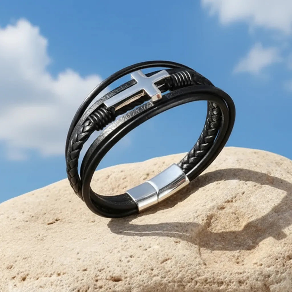 Bracelet multi-rangs en cuir noir dressé et penché sur un rocher, avec vue sur la croix latine et sur le fermoir sur fond de ciel bleu.