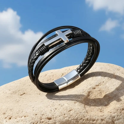 Bracelet multi-rangs en cuir noir dressé et penché sur un rocher, avec vue sur la croix latine et sur le fermoir sur fond de ciel bleu.