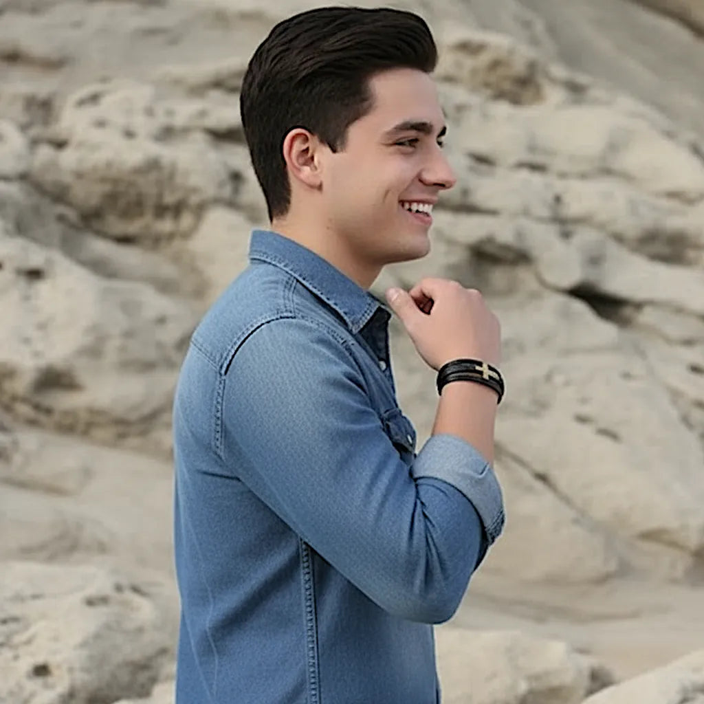 Jeune homme souriant de profil, portant un bracelet en cuir noir avec croix latine argentée sur fond rocheux.