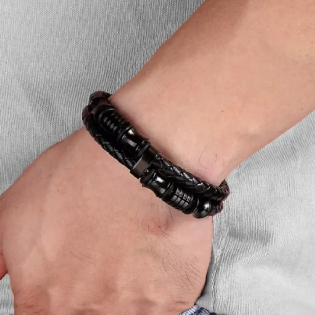 Bracelet en cuir noir tressé, porté sur la main gauche d’un homme. Le contraste des perles diabolo et des cylindres en acier inoxydable brillant rehausse l’élégance du bijou. Accessoirisé par une tenue avec sweat gris clair pour un look casual chic.