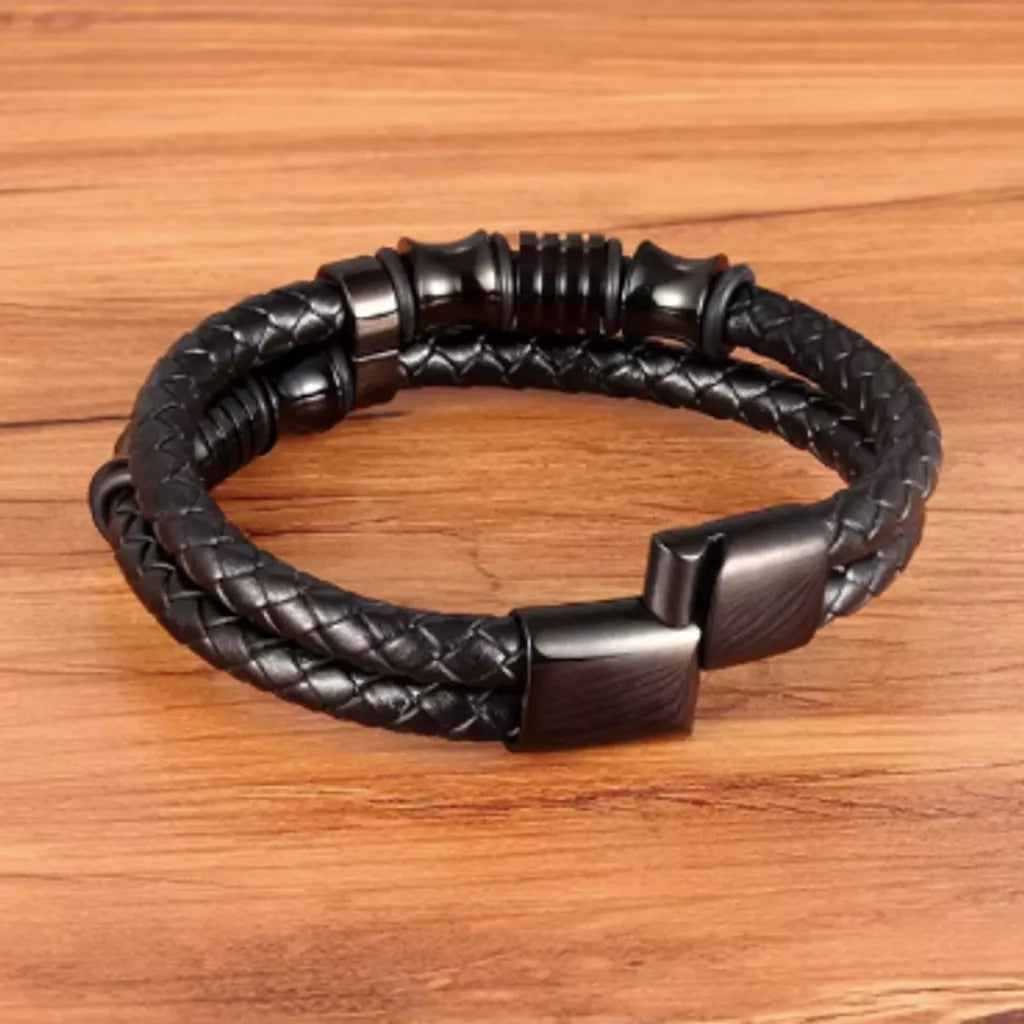 Bracelet en cuir tressé noir, posé à plat sur une surface en bois. Le fermoir noir magnétique est entrouvert, offrant un aperçu des finitions robustes. Les perles diabolo et cylindres noirs mettent en avant un style audacieux et contemporain.