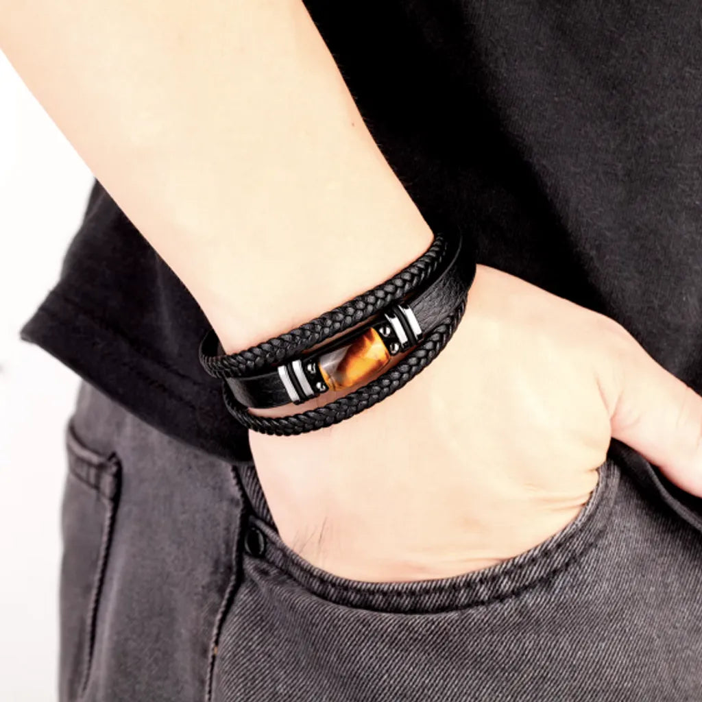 Bracelet en cuir noir avec double tresse et œil de tigre jaune, porté sur le poignet droit d'un homme. Ce bracelet à fermoir magnétique argenté ajoute une touche de caractère et d'élégance au look, parfait pour compléter un style masculin.