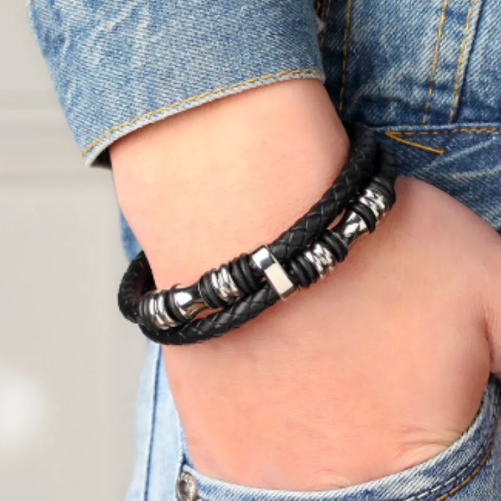 Bracelet en cuir noir tressé porté sur un poignet masculin. Le design intègre des perles diabolo et des cylindres en acier inoxydable brillant. L’ensemble est associé à une tenue en jean décontractée, mettant en valeur le style moderne et intemporel du bracelet.