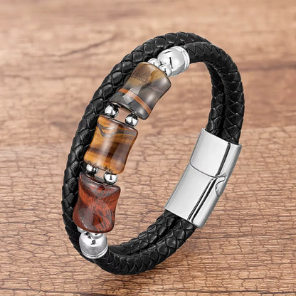 Bracelet noir en cuir tressé, décoré de pierres œil de tigre jaune, rouge et bleu, formant un contraste vibrant avec le fermoir magnétique en acier argenté. La scène est capturée sur un arrière-plan en bois patiné, évoquant une ambiance artisanale et authentique.