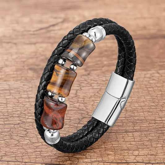 Bracelet noir en cuir tressé, décoré de pierres œil de tigre jaune, rouge et bleu, formant un contraste vibrant avec le fermoir magnétique en acier argenté. La scène est capturée sur un arrière-plan en bois patiné, évoquant une ambiance artisanale et authentique.