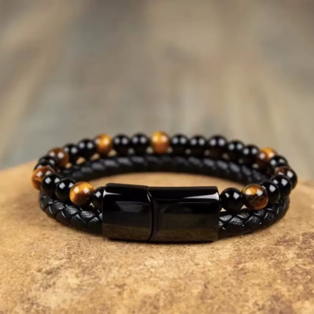 Bracelet double rang en cuir tressé noir, agrémenté de perles rondes d’œil de tigre jaune doré et de perles d’onyx noir profond. Présenté sur une surface rocheuse texturée dans des teintes naturelles beige et marron, avec un arrière-plan flou aux nuances terreuses, l’image met en avant le fermoir magnétique noir brillant et l’élégance intemporelle du design. Une composition idéale pour refléter un style masculin chic.