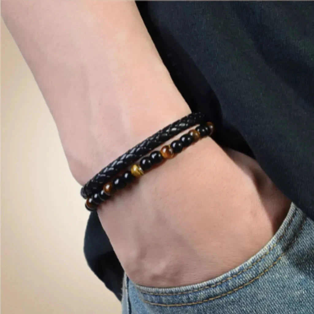 Bracelet double rang en cuir tressé noir, orné de perles d’œil de tigre jaune doré et d’onyx noire, porté sur la main droite. Le design raffiné et les teintes naturelles des pierres apportent une touche élégante et masculine. L’image montre un poignet vêtu de denim et d’un t-shirt noir, dans une ambiance minimaliste avec un fond beige doux.