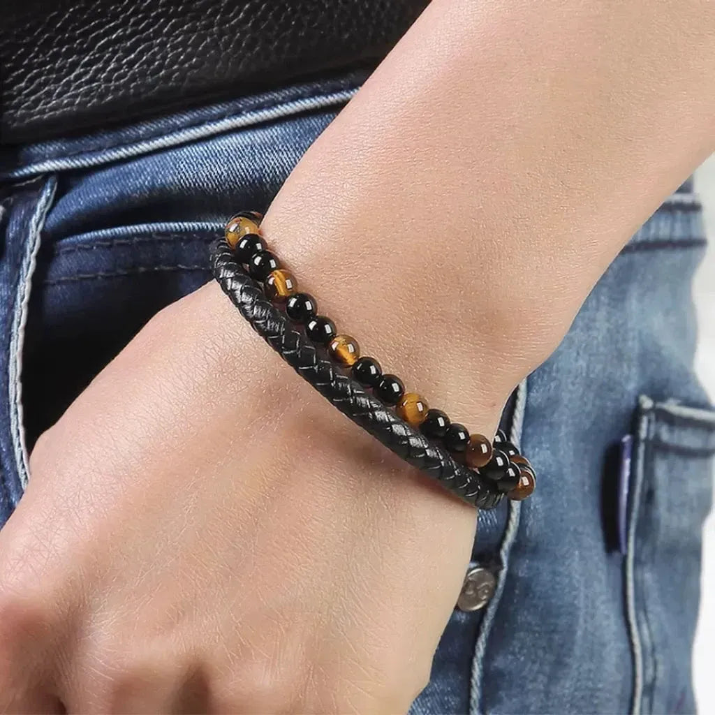 Bracelet en cuir tressé noir agrémenté de perles de pierre jaune lumineuses et d’onyx noir, porté sur la main gauche. L’image présente un look décontracté, un pantalon en jean bleu subtil qui met en valeur les nuances éclatantes des pierres et l’élégance moderne du bracelet.