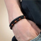 Bracelet en cuir tressé noir avec des perles de pierre rouge profond et des accents d’onyx noire, porté sur un poignet masculin droit. L’ensemble est présenté dans un style décontracté avec un jean et un t-shirt noir, le fond beige adoucit la composition et met en valeur l’élégance des pierres.