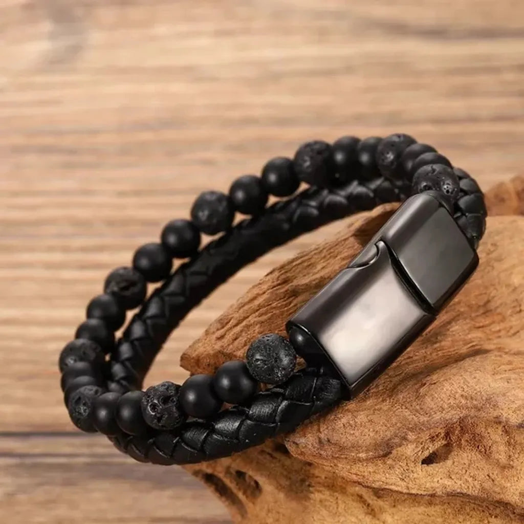 Un bracelet en onyx noir et pierre de lave naturelle, posé élégamment sur un socle en bois texturé. Les détails mettent en valeur les perles mates et la lanière en cuir tressé, accompagnés d'un fermoir magnétique, métallique et brillant, idéal pour un look naturel et moderne.