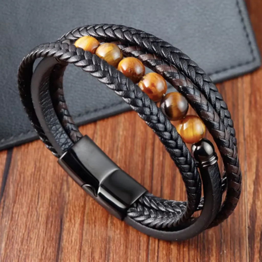 Bracelet en cuir tressé noir et marron, orné de perles d’œil de tigre jaune scintillant, présenté dans une vue, où ce wrap est dressé et de trois quarts de face. Le bijou repose sur une table en bois, accompagné d’un portefeuille en cuir noir qui ajoute une touche de sophistication au décor.