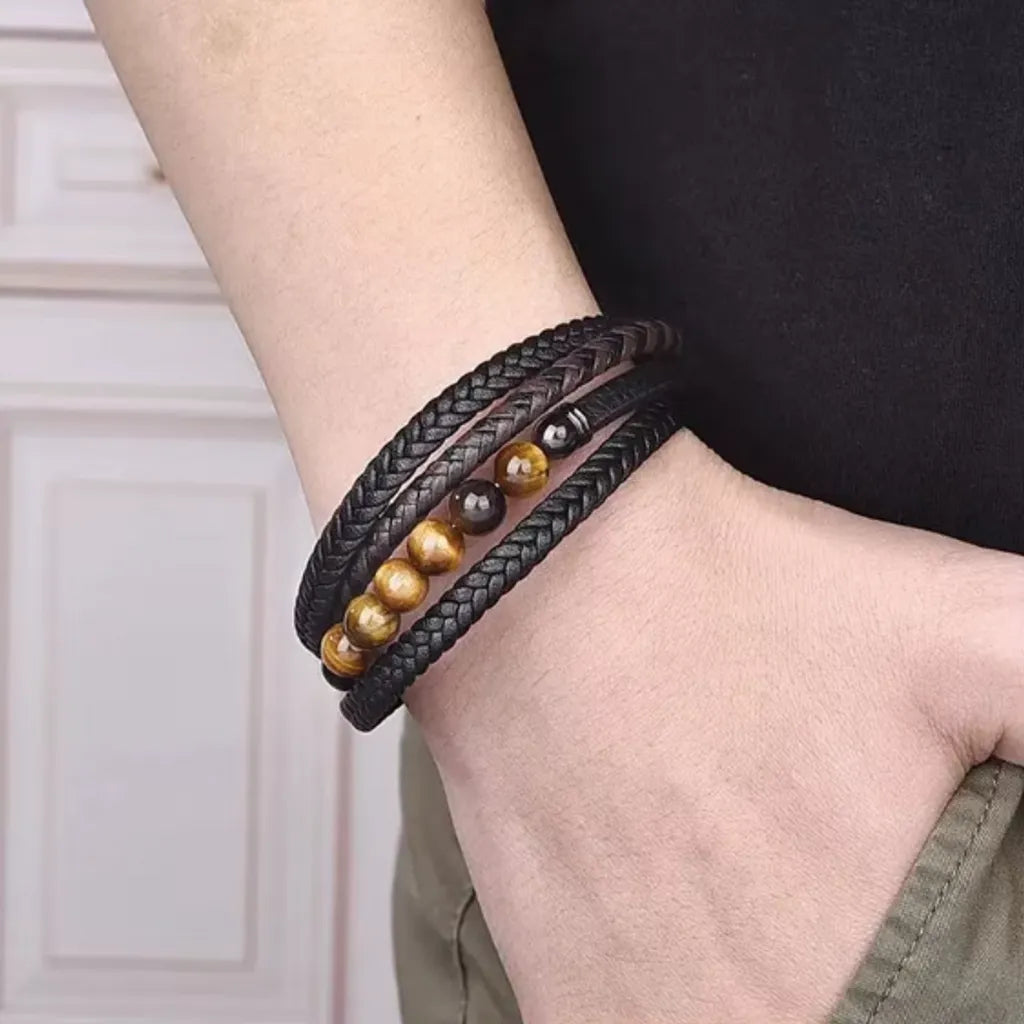 Bracelet multitours en cuir tressé noir et marron, orné de perles d’œil de tigre jaune brillant et d'accents métallique noir. Porté au poignet masculin, il offre une allure élégante et décontractée, idéal pour compléter une tenue moderne avec un look chic et affirmé.