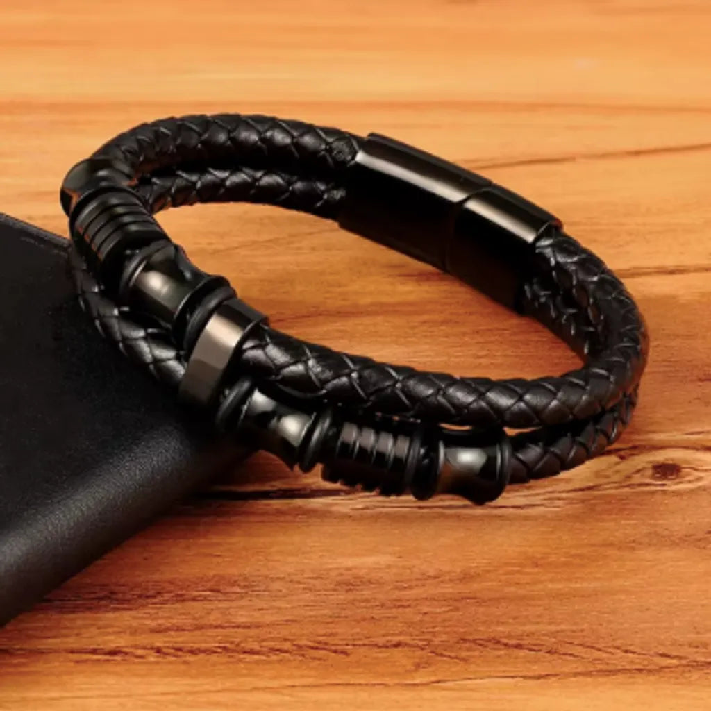 Bracelet en cuir tressé noir avec des perles diabolo et cylindres noirs, présenté sur un fond en bois naturel pour un effet chaleureux. Le bracelet repose élégamment sur un portefeuille en cuir noir, soulignant son design moderne et masculin.