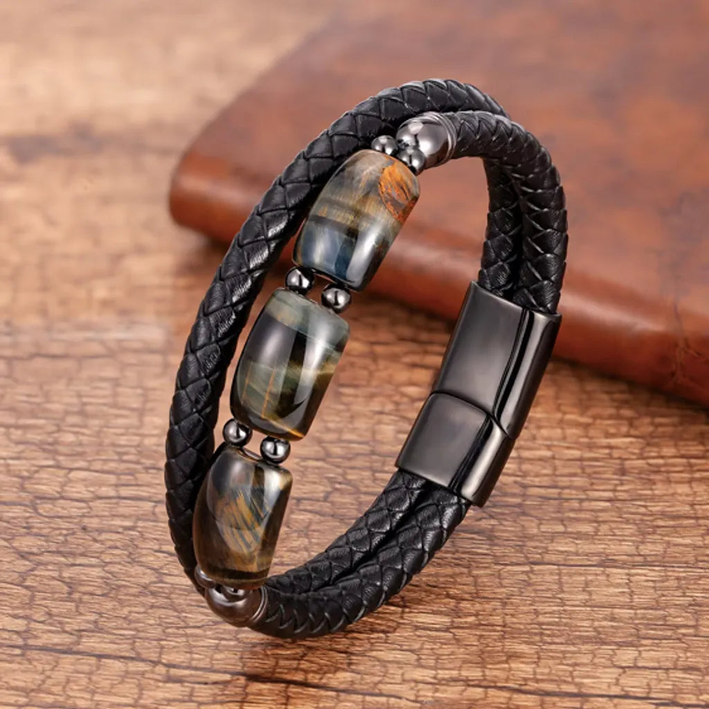 Bracelet en cuir noir tressé avec trois pierres œil de tigre bleu profond, entourées de perles et d’accents en acier noir mat. Le bijou repose sur une surface en bois rustique, apportant un contraste naturel et chaleureux.