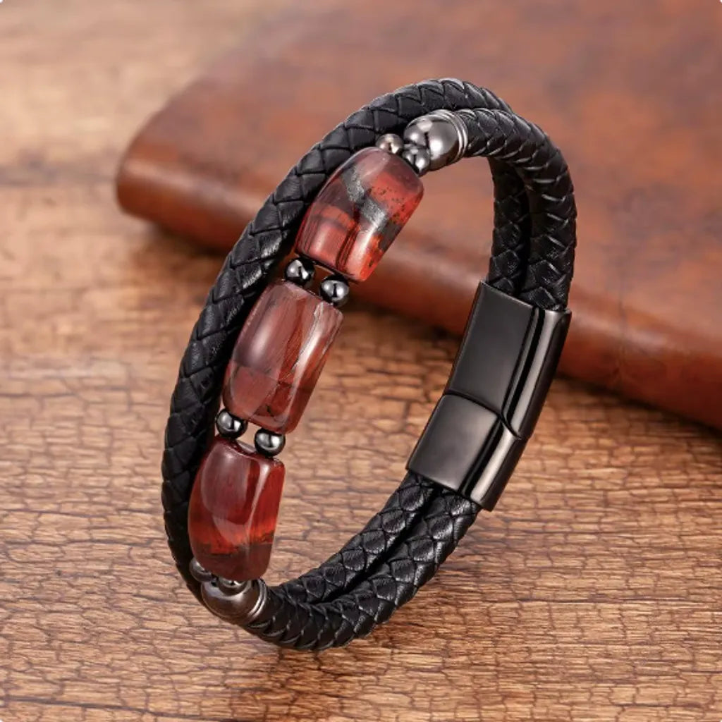 Bracelet noir tressé en cuir de qualité, agrémenté de trois pierres œil de tigre rouge au fini brillant, présenté sur un fond en bois brun au grain marqué. Une pièce tendance pour un style audacieux et affirmé.