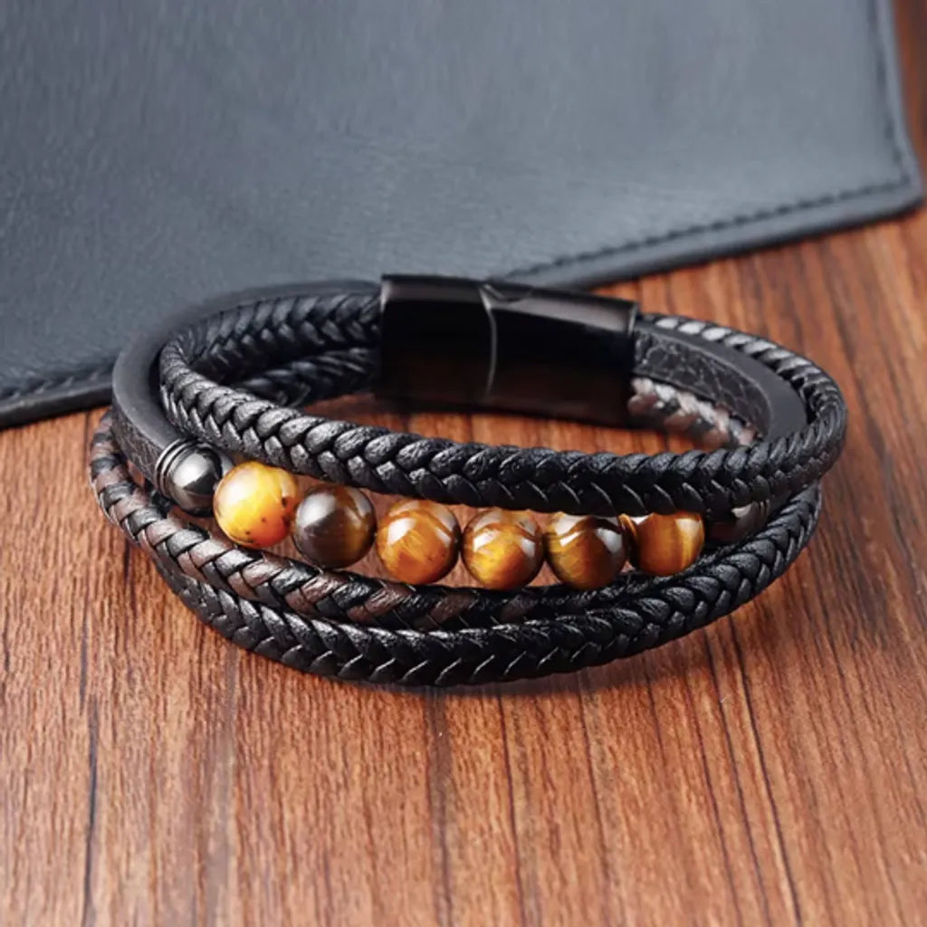 Bracelet en cuir tressé noir avec une touche de marron, présenté à plat avec une vue centrée sur les perles d’œil de tigre jaune, parfaitement alignées. Le fermoir en acier inoxydable noir est fermé, ajoutant une touche de finition au design. Le décor comprend une table en bois chaleureuse et un portefeuille en cuir noir pour un style intemporel.