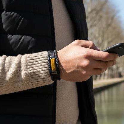 Focus sur le Bracelet Fauve porté au poignet, révélant le contraste entre les tresses noires et l'œil de tigre solaire. L'accessoire ponctue une tenue casual chic devant les eaux paisibles du Canal du Midi.