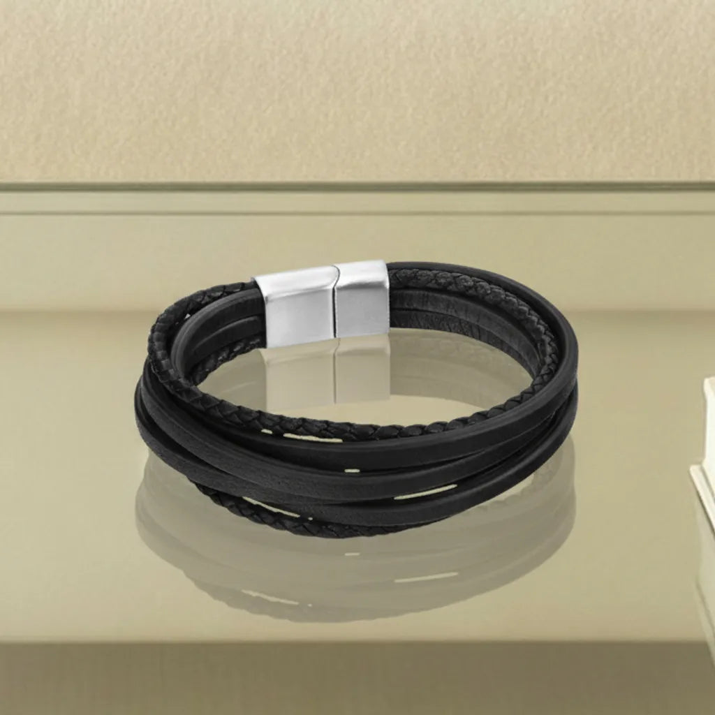 Vue détaillée. Le bracelet Fusion cuir souple noir posé à plat, révélant le mélange des six lanières tressées et lisses. Le fermoir en acier inoxydable capte la lumière sur le verre.