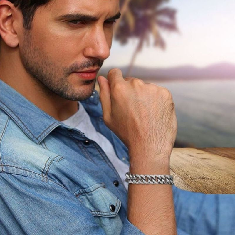 Bracelet gourmette à maillons cubains de quinze millimètres avec fermoir sécurisé en acier inoxydable de couleur argent porté par un jeune homme pensif au bord de la mer