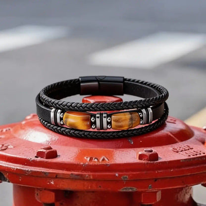 Composition horizontale du bracelet multi-rangs Apex posé sur une surface métallique rouge, où la bande de cuir centrale et ses gemmes d'œil de tigre contrastent avec la texture brute de l'acier urbain.